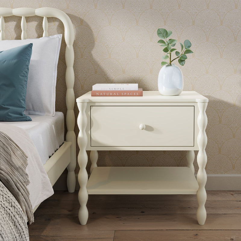 Cream spindle-leg bedside table nightstand with single drawer and lower shelf, white ceramic vase with eucalyptus, stacked books, teal pillow, textured beige wallpaper and wood floor, cozy modern farmhouse bedroom decor