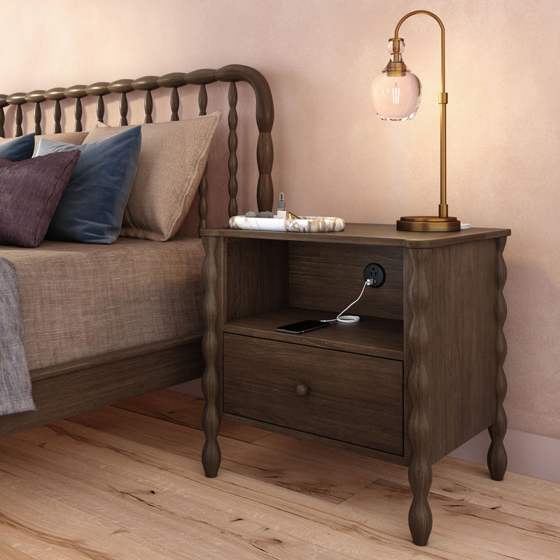 Dark wood bedside table with carved legs, built-in power outlet and charging smartphone, brass lamp and decorative tray next to upholstered bed on hardwood floor