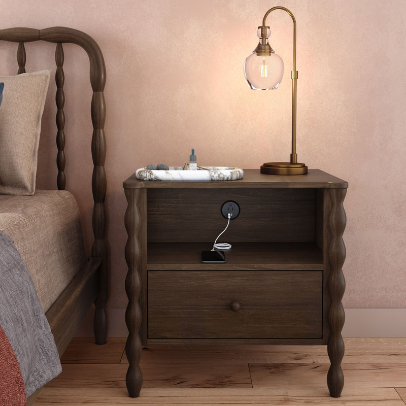 Dark wooden nightstand with built-in power outlet and USB charger, smartphone charging, brass table lamp and marble tray beside spindle bed against pink textured wall