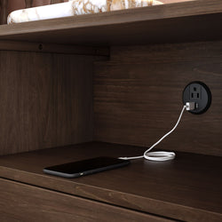 Smartphone charging on a wooden nightstand shelf using a round built-in USB power outlet — bedside charging station with tidy cord management.