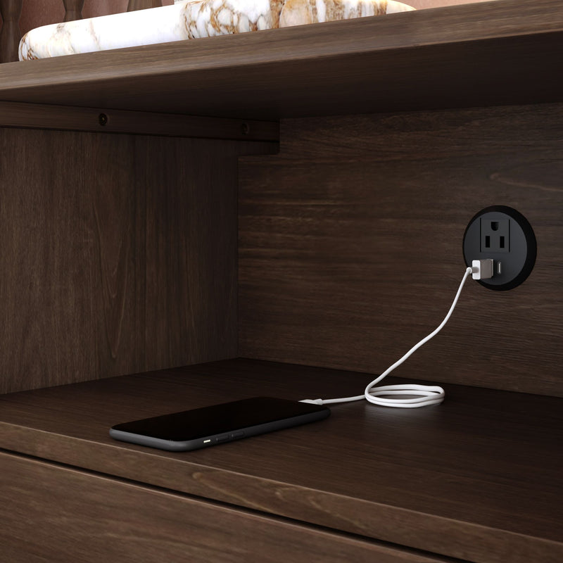 Smartphone charging on a wooden nightstand shelf using a round built-in USB power outlet — bedside charging station with tidy cord management.