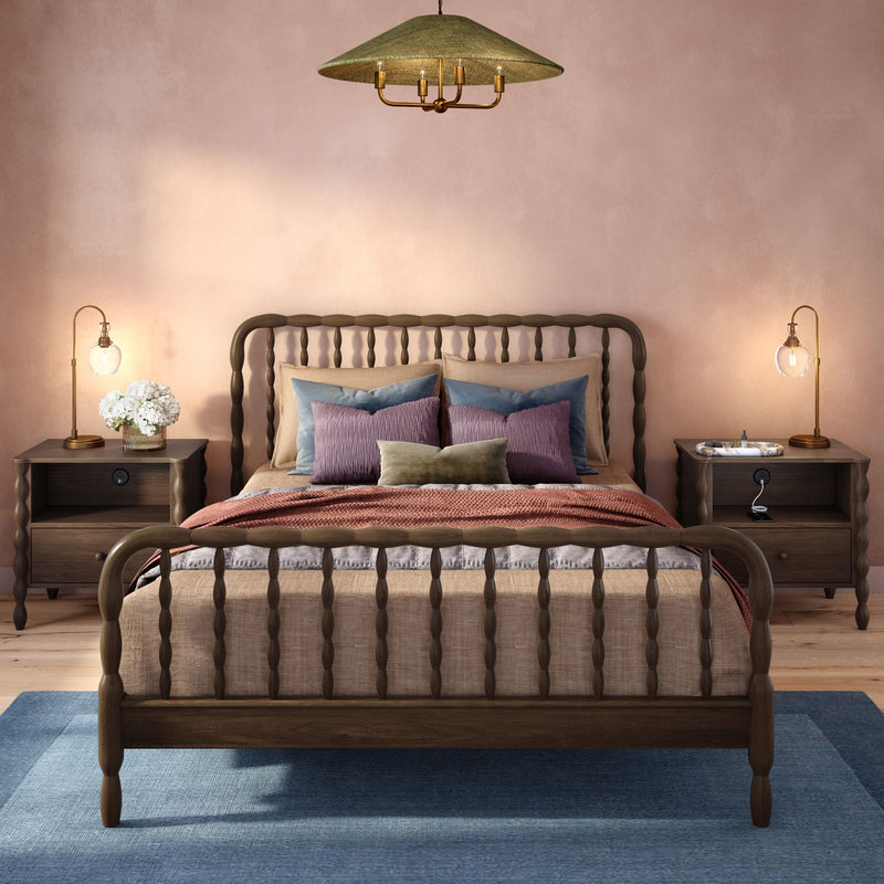 Cozy modern farmhouse bedroom with carved wooden spindle bed, blush pink textured wall, layered neutral and rust bedding with blue and mauve throw pillows, matching dark wood nightstands with brass table lamps, blue area rug and pendant light