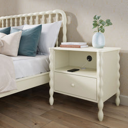 Cream bedside table with turned legs and built-in charging outlet, styled with vase, books and smartphone beside a matching spindle bed on hardwood floor