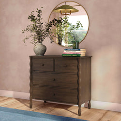 Dark wood four-drawer dresser with carved legs topped by a round gold mirror, vase of greenery and stacked books against a soft pink wall on a wooden floor
