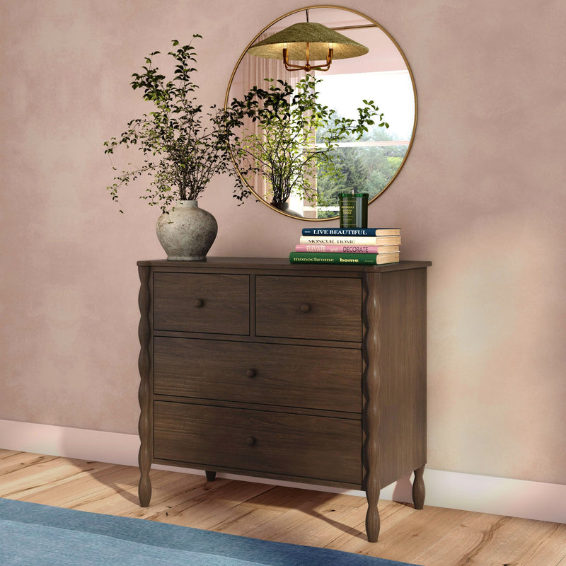 Dark wood four-drawer dresser with carved legs topped by a round gold mirror, vase of greenery and stacked books against a soft pink wall on a wooden floor
