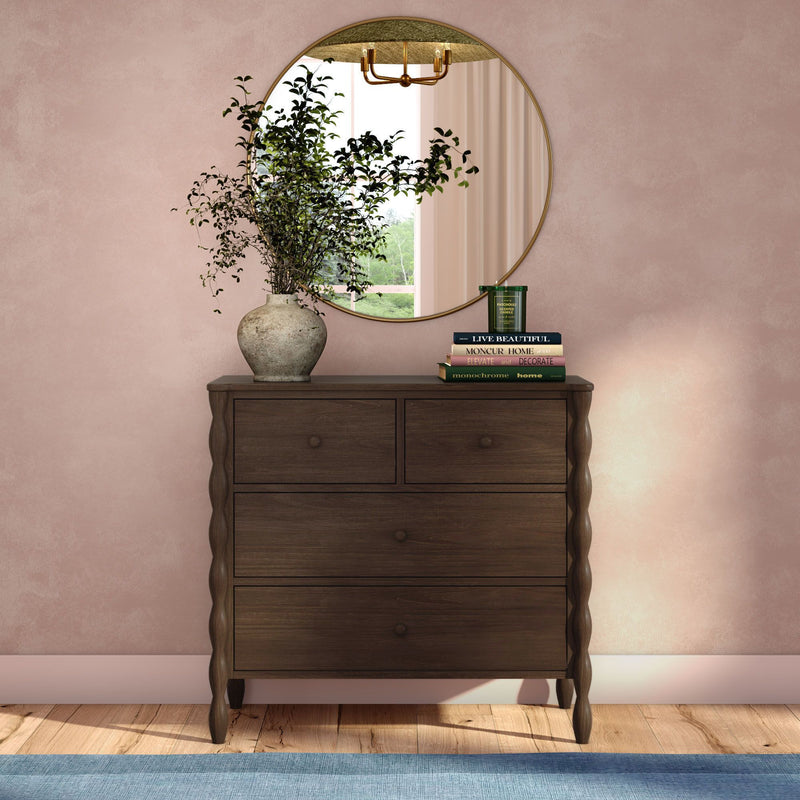 Dark wood scalloped-leg dresser in a modern farmhouse entryway with a round gold-framed mirror, rustic stone vase of green foliage, stacked home-decor books and candle against a blush pink textured wall over hardwood floor and blue rug