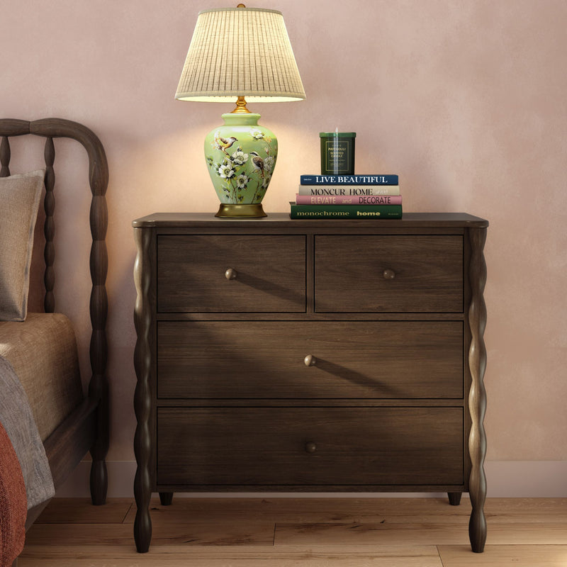 Dark wood nightstand with twisted legs, green floral ceramic table lamp, stacked home decor books and scented candle on hardwood floor against a blush wall