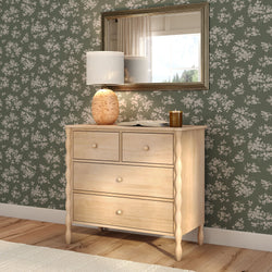 Light wood dresser with turned legs and drawers against green floral wallpaper, ceramic table lamp and gold-framed mirror in cozy bedroom decor.