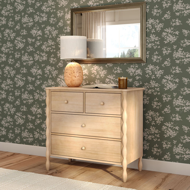 Light wood dresser with turned legs and drawers against green floral wallpaper, ceramic table lamp and gold-framed mirror in cozy bedroom decor.