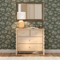 Light wood four-drawer dresser with ceramic table lamp and framed mirror against green floral wallpaper — cozy bedroom decor and rustic modern interior styling