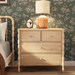 Light wood bedroom dresser with turned legs and drawers, glowing ceramic table lamp, oval framed photo and green floral wallpaper