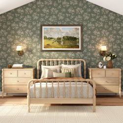 Cozy farmhouse bedroom with light wood spindle bed, green floral wallpaper, neutral patterned bedding and throw pillows, matching wooden nightstands, wall sconces, framed landscape painting and vase of white flowers.