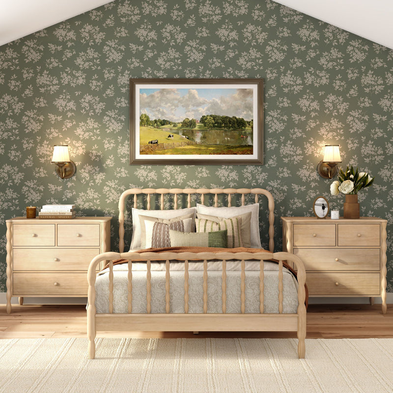 Cozy farmhouse bedroom with light wood spindle bed, green floral wallpaper, neutral patterned bedding and throw pillows, matching wooden nightstands, wall sconces, framed landscape painting and vase of white flowers.