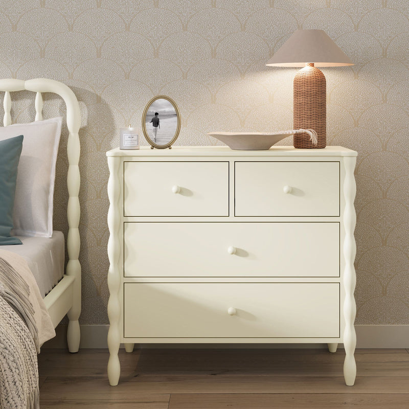 Cream wooden four-drawer bedside dresser with rattan table lamp, decorative bowl, framed beach photo and candle on hardwood floor against beige patterned wallpaper in a neutral bedroom
