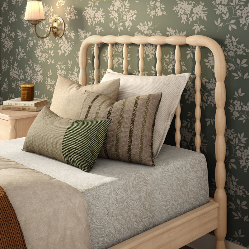 Cozy cottage-style bedroom with light wooden spindle bed, green floral wallpaper, layered neutral and striped throw pillows, textured paisley bedding, bedside table with candle and books, and brass wall sconce