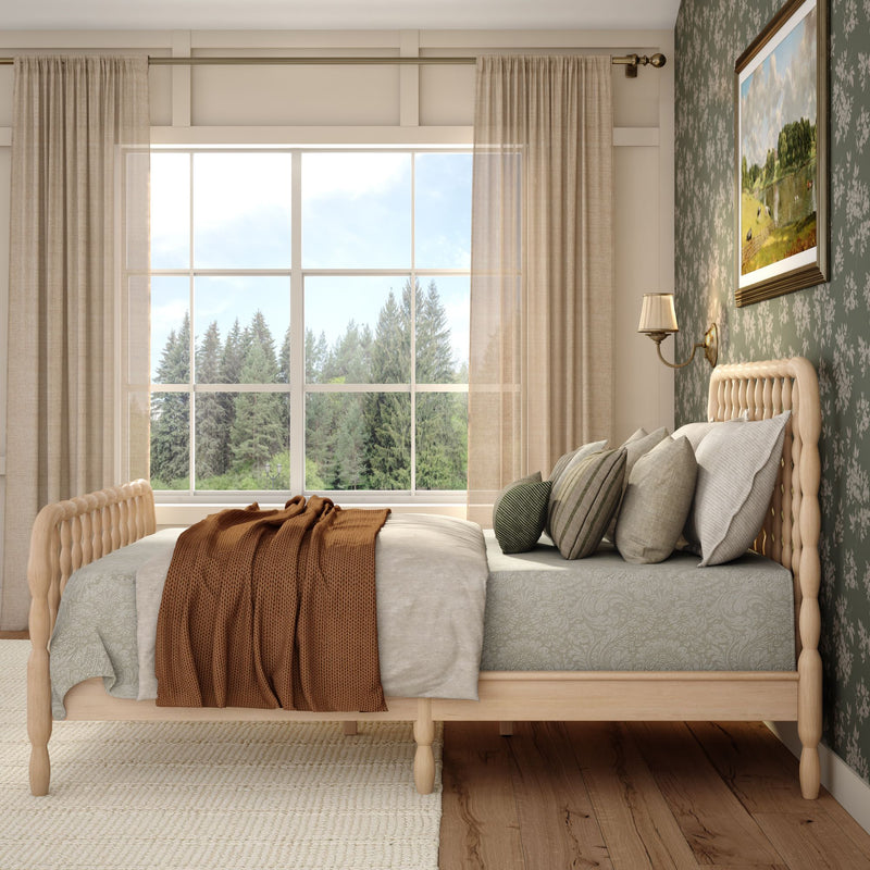Cozy modern farmhouse bedroom with light wooden spindle bed, neutral linen bedding and rust knit throw, layered pillows, floral green accent wall, sheer curtains and large window with pine forest view