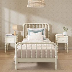 Cream spindle twin bed with layered pillows and beige bedding flanked by matching nightstands in a cozy neutral bedroom with patterned wallpaper, wood floors, lamp, plant and rattan pendant light