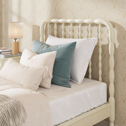 Cozy bedroom with cream spindle headboard, beige patterned wallpaper, neutral linen bedding, teal accent pillow and wooden bedside lamp