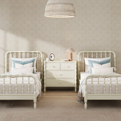 Neutral children's bedroom with two cream spindle twin beds, blue accent pillows, cream three-drawer dresser, table lamp and wicker pendant light