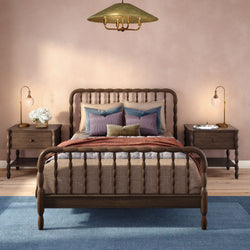 Cozy modern farmhouse bedroom with wooden spindle bed, layered neutral bedding and decorative pillows, blush-pink wall, blue area rug, matching wooden nightstands and brass bedside lamps