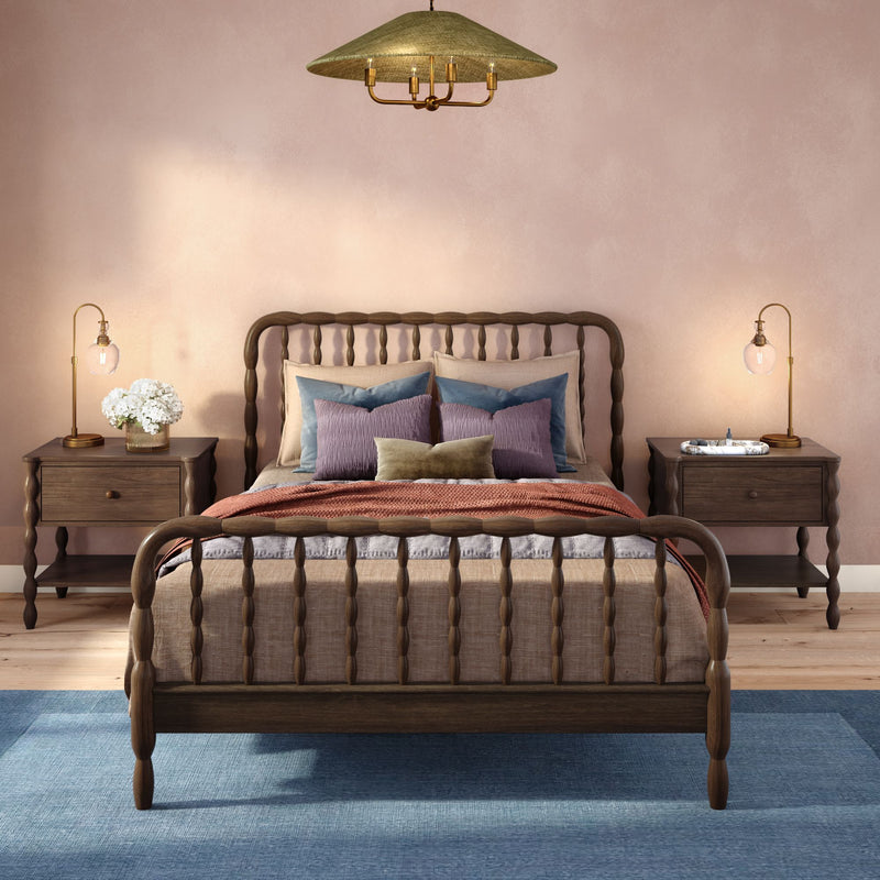 Cozy modern farmhouse bedroom with wooden spindle bed, layered neutral bedding and decorative pillows, blush-pink wall, blue area rug, matching wooden nightstands and brass bedside lamps