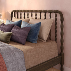 Cozy bedroom with dark wood spindle headboard, neutral linen bedding and textured velvet decorative pillows in purple, blue, and olive against a blush pink wall