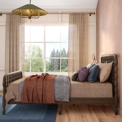 Cozy modern bedroom interior with carved wooden bed frame, neutral linen bedding and rust throw blanket, decorative pillows, large window with sheer curtains and forest view, pendant light and hardwood floors.