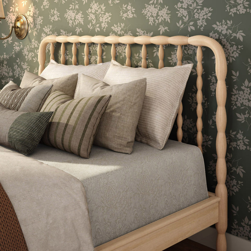 Cozy farmhouse bedroom decor featuring a light wooden spindle headboard, green floral wallpaper, layered neutral linen and striped accent pillows, textured beige duvet, and brass wall sconce