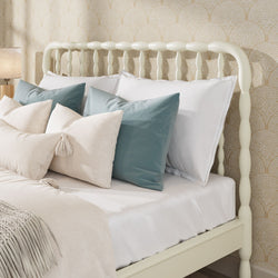 Cream spindle bed frame with layered white and teal throw pillows, neutral linen bedding and tasseled accent pillows against gold textured wallpaper