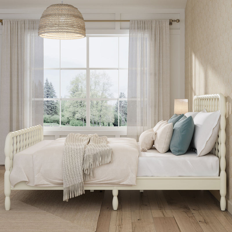 Cozy neutral farmhouse bedroom with white spindle bed, layered pillows and knit throw, large window with sheer curtains, rattan pendant light and light hardwood floors