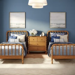 Coastal twin bedroom with navy blue accent wall, wooden twin beds, blue pillows, wood nightstand with white flowers and ocean wall art
