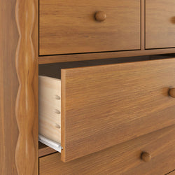Close-up of a wooden bedroom dresser with a partially open drawer showing warm natural wood grain, round wooden knob, visible drawer joinery and metal slide