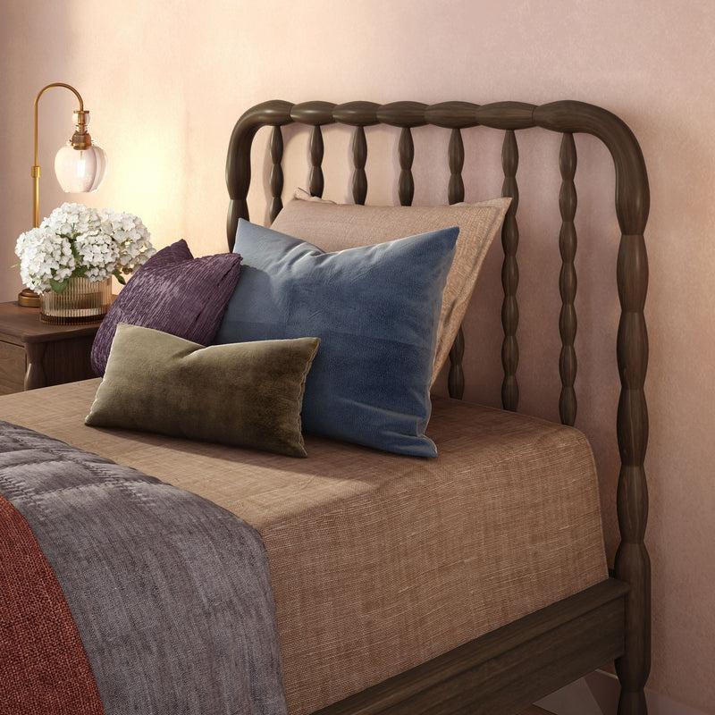 Cozy bedroom close-up with dark wooden spindle headboard, layered velvet and linen pillows in blue, purple and olive, beige bedding, gold bedside lamp and glass vase of white hydrangeas on a wooden nightstand against a soft pink wall, modern neutral interior decor