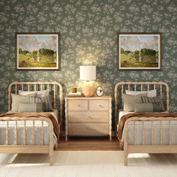 Cozy cottage-style twin bedroom with light wood spindle headboards, neutral layered bedding and pillows, green floral wallpaper, framed landscape paintings, wooden nightstand with textured lamp and farmhouse decor