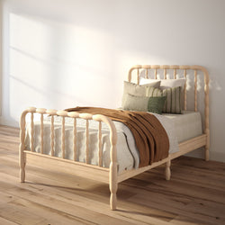 Sunlit minimalist bedroom with spindle wood bed, neutral bedding, green-striped pillows and brown throw on hardwood floor.