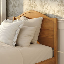 Wooden arched bed headboard with beige linen bedding and ruffled pillows against floral wallpaper in a cozy farmhouse bedroom