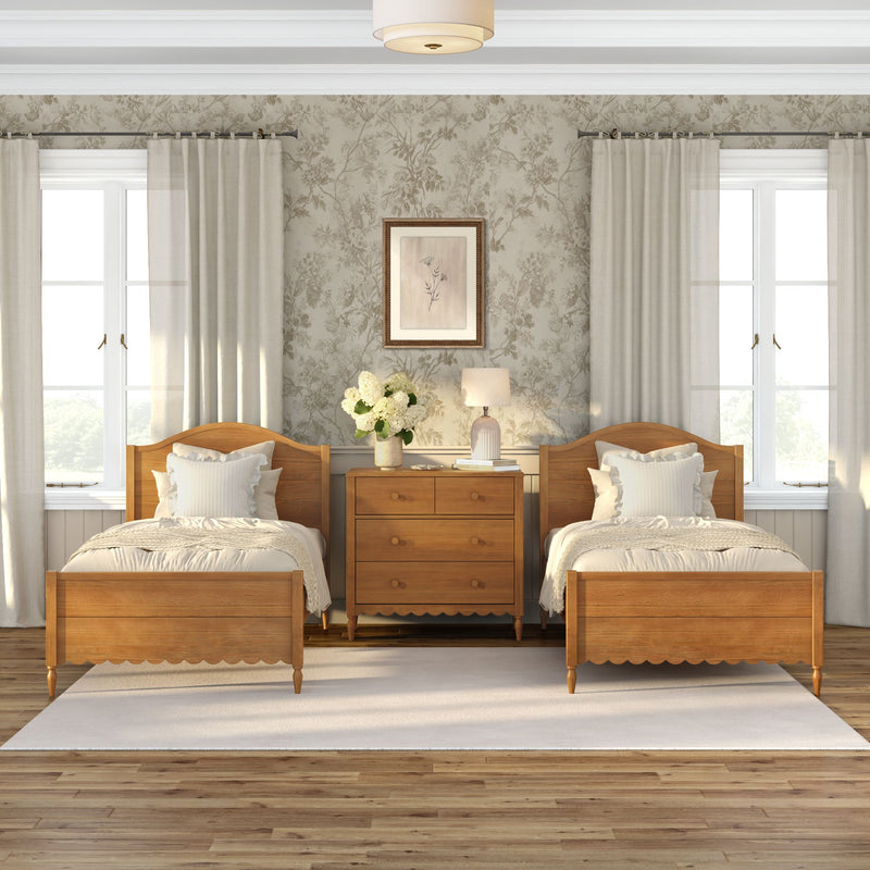Cozy farmhouse-style twin bedroom interior with matching wooden beds, central wooden dresser, neutral floral wallpaper, white curtains, bedside lamp and vase of white hydrangeas.