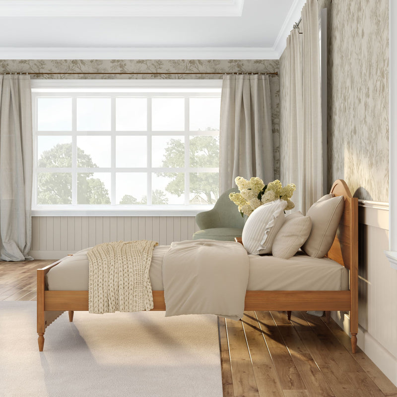 Cozy neutral bedroom with wooden bed frame, beige bedding and knit throw, large grid window with sheer curtains and natural light, green accent chair with white flower bouquet, floral wallpaper and hardwood floors