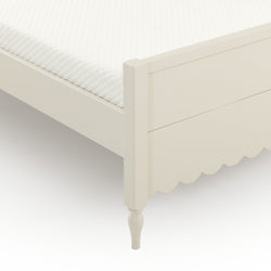 Close-up of cream white wooden bed frame corner with quilted mattress topper, scalloped footboard and turned leg