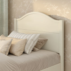 Cream curved wooden headboard with neutral beige bedding and striped and gingham pillows against beige floral wallpaper, cozy farmhouse bedroom decor