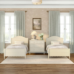 Neutral twin bedroom with cream scalloped beds, white dresser, lamp and hydrangea vase, pale green curtains and hardwood floor