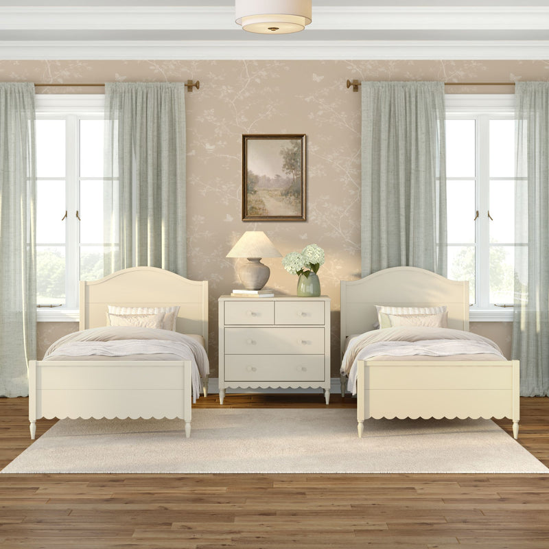 Neutral twin bedroom with cream scalloped beds, white dresser, lamp and hydrangea vase, pale green curtains and hardwood floor