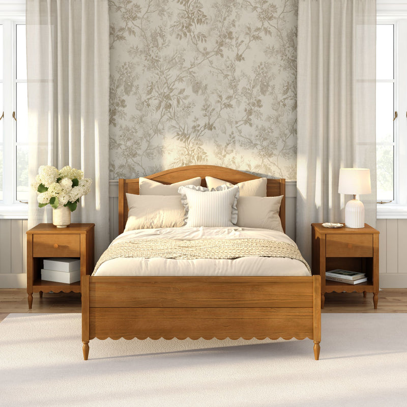Cozy farmhouse bedroom with wooden bed frame, neutral bedding and throw, floral wallpaper, matching nightstands, lamp, white hydrangea vase and soft natural light
