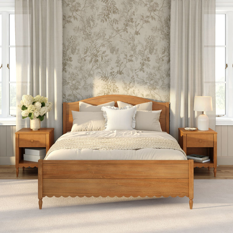 Sunlit neutral farmhouse bedroom with wooden bed frame, cream bedding and knit throw, floral wallpaper accent wall, matching wooden nightstands, table lamp, vase of white hydrangeas and sheer curtains