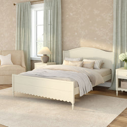 Cozy neutral farmhouse bedroom interior with a cream wooden bed and scalloped footboard, layered beige and gray bedding, light green curtains, beige floral wallpaper, upholstered armchair, matching nightstands with lamp and vase of white flowers, and a soft area rug on hardwood floors