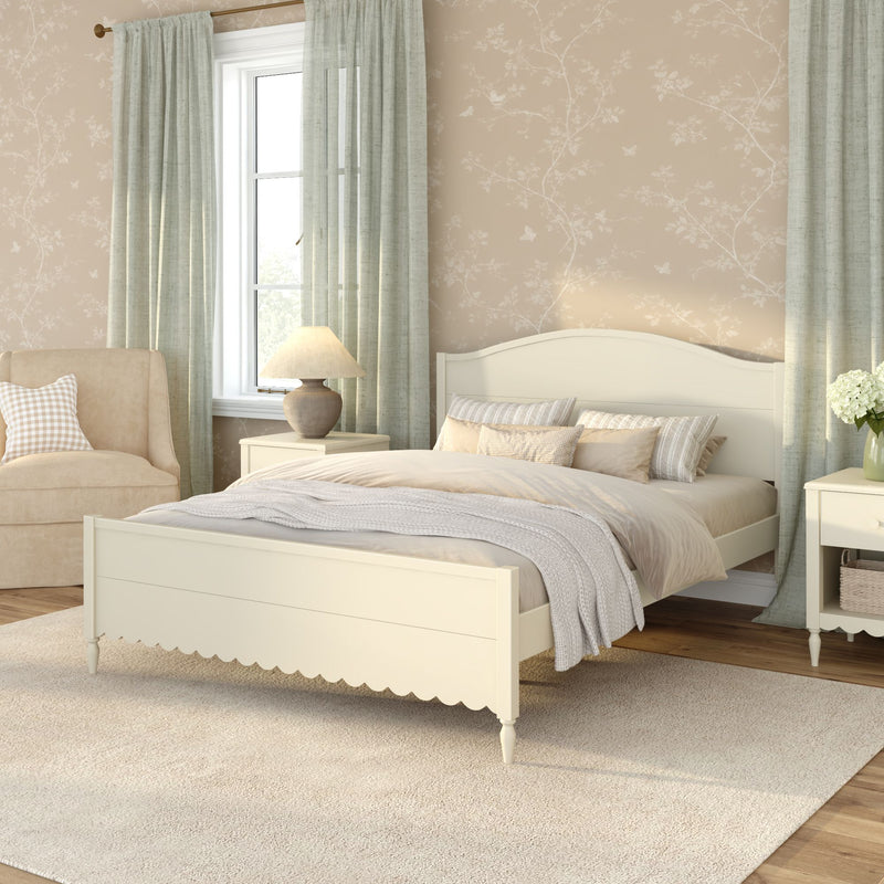 Cozy neutral farmhouse bedroom interior with a cream wooden bed and scalloped footboard, layered beige and gray bedding, light green curtains, beige floral wallpaper, upholstered armchair, matching nightstands with lamp and vase of white flowers, and a soft area rug on hardwood floors
