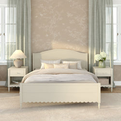 Cozy neutral farmhouse bedroom with cream wooden bed, beige floral wallpaper, matching nightstands with lamps, sage green curtains, soft layered bedding and vase of white hydrangeas by bright windows