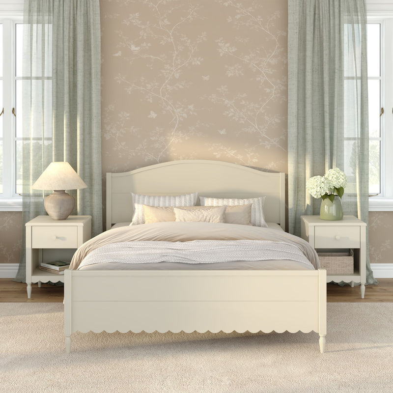 Cozy neutral farmhouse bedroom with cream wooden bed, beige floral wallpaper, matching nightstands with lamps, sage green curtains, soft layered bedding and vase of white hydrangeas by bright windows