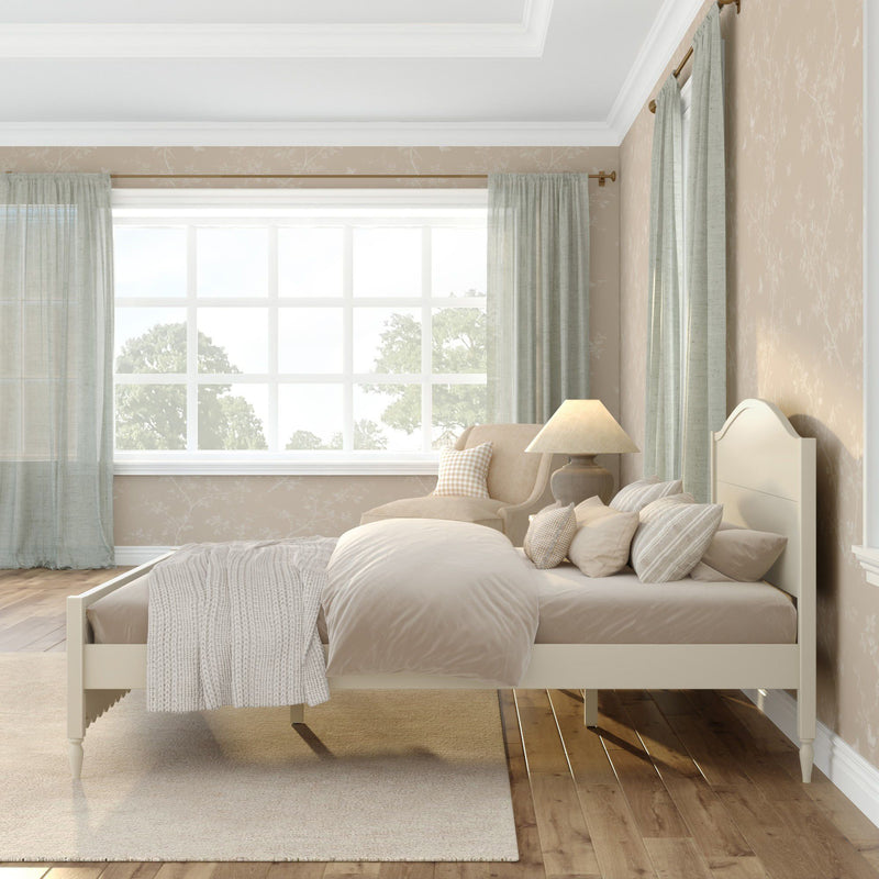 Cozy sunlit neutral bedroom interior with beige bedding and knitted throw, decorative pillows, upholstered armchair, bedside lamp, light-green curtains, and hardwood floors