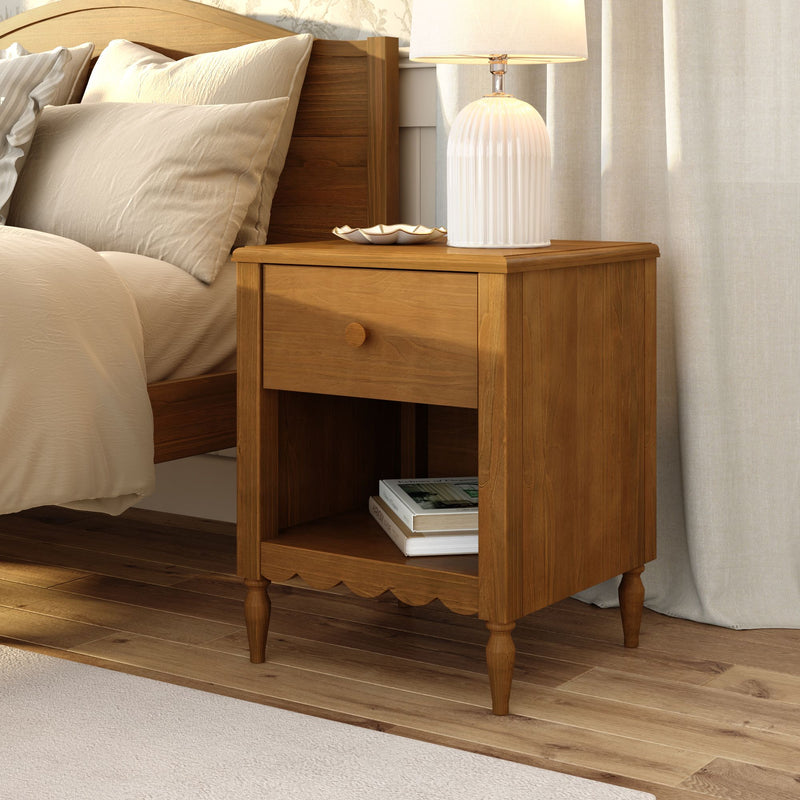 Warm wooden nightstand with drawer and white ceramic table lamp next to beige linen pillows on a bed in a cozy modern farmhouse bedroom